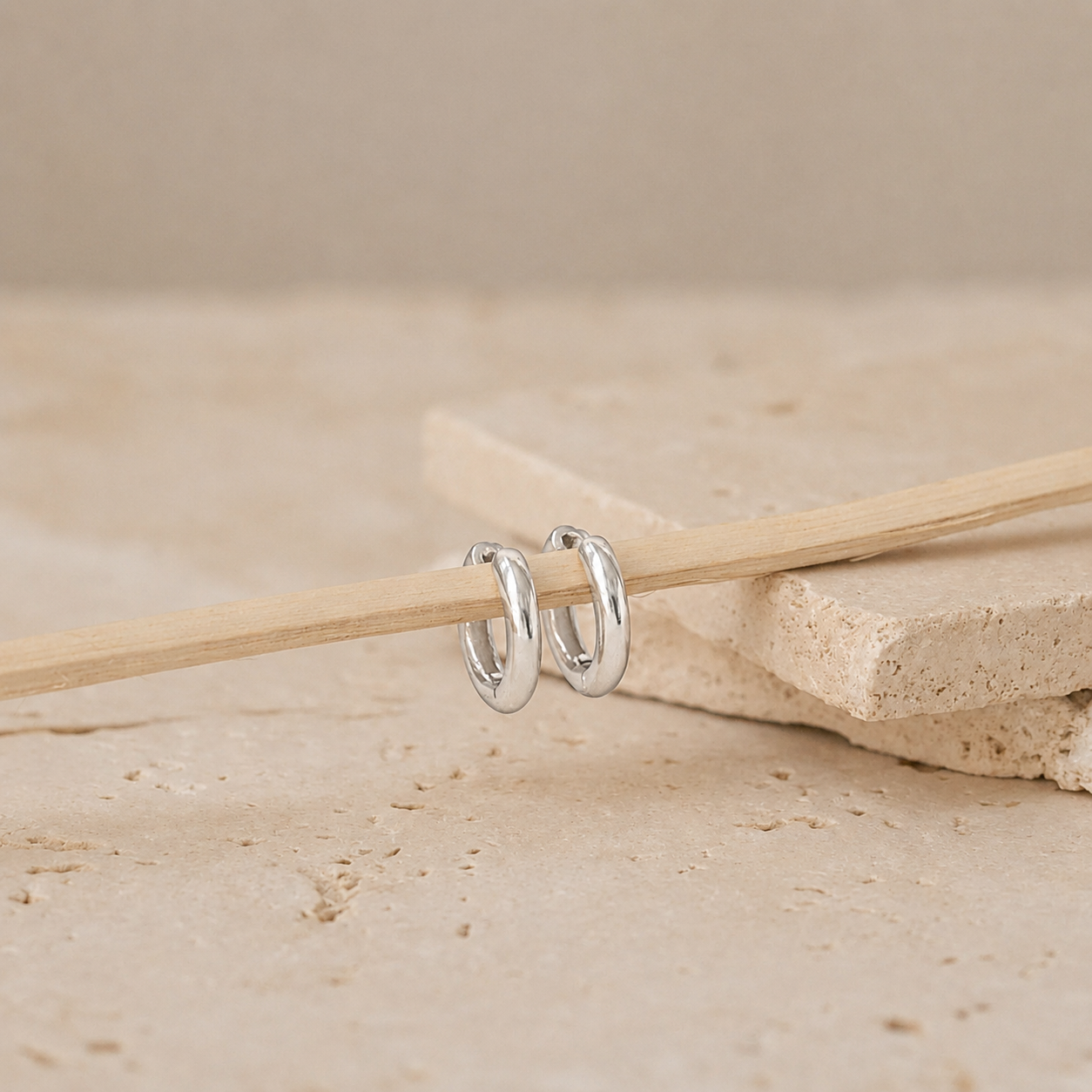 A pair of polished silver hoop earrings resting on a light wooden stick, set against a neutral beige stone background.